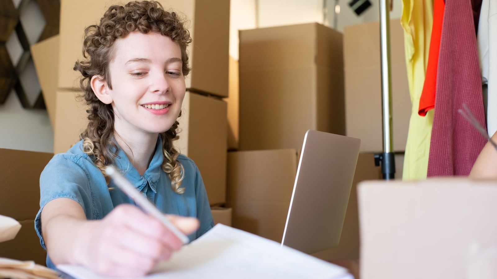 A smiling small business owner writing notes at her desk, surrounded by inventory boxes