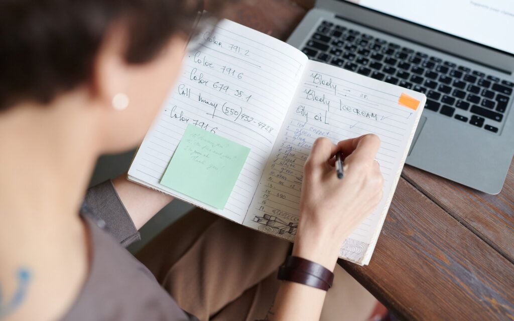 A person writing a short numbered outline in a notebook next to a laptop