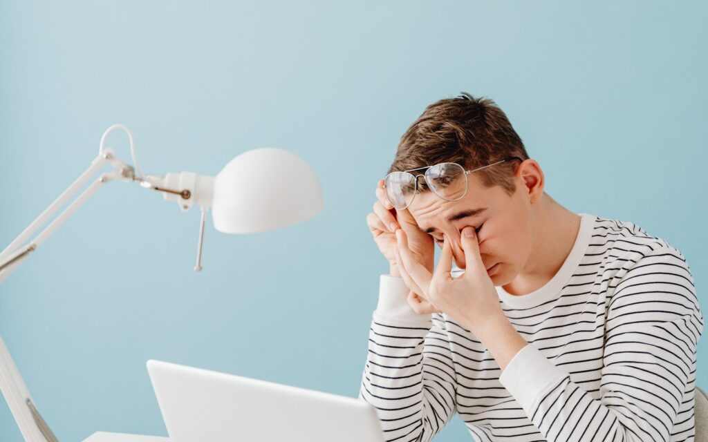 A tired person rubbing their eyes at a desk in the evening with a lamp on and a laptop open