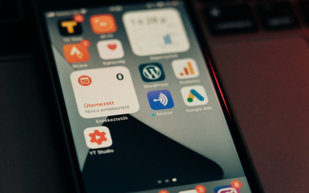 A phone home screen full of work and marketing apps, held next to an open laptop