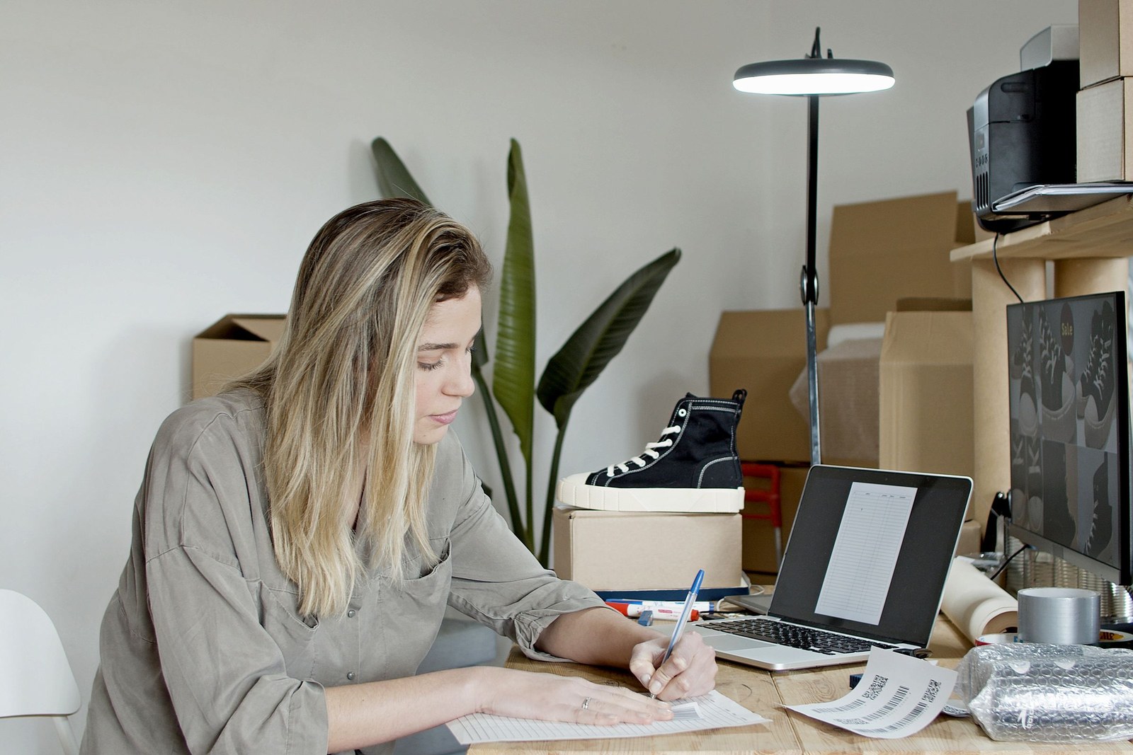 A woman writing at a desk alone in a small office with boxes and a plant behind her