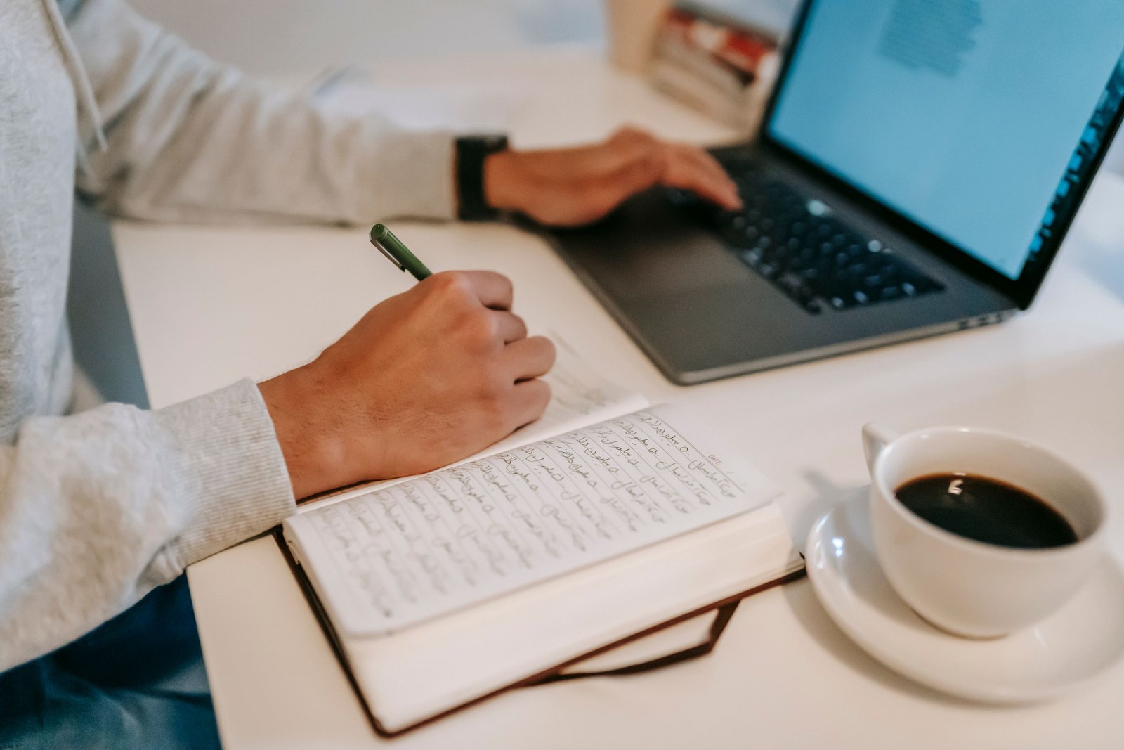 A person writing notes in a notebook next to an open laptop and a cup of coffee